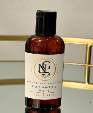 A bottle of Noerf Gold Skin Cashmere Veil Nourishing Body Oil, featuring an amber glass bottle, a black cap, and a cream-colored label with gold accents, set against a softly blurred background with a gold metallic frame.